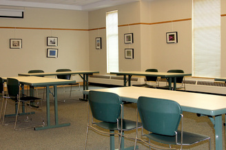 Photo of Main Meeting Room. Carpeted meeting room with four tables; each table has two chairs placed next to it. Small, framed art pieces and photographs are hung on the walls. Two tall windows are on the far wall.