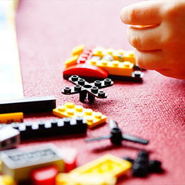 A small hand holds a LEGO piece above a LEGO structure with other pieces scattered around the floor..